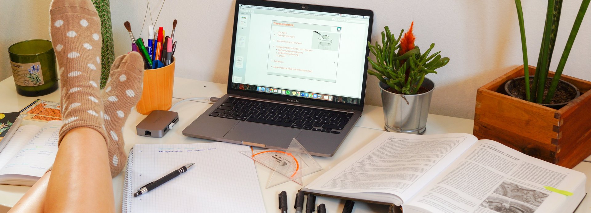 Schreibtisch mit Laptop, Lernunterlagen und Dekoration. Ein Studierender hat die Füße auf dem Schreibtisch abgelegt.
