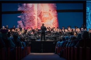 Das Universitätsblasorchester bei einem Konzert auf der Bühne. Der Dirigent steht mit dem Rücken zum Publikum.