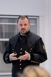 Eine Person in schwarzer bergmännischer Festuniform mit goldenen Knöpfen und Abzeichen hält einen Vortrag in einem Innenraum und gestikuliert.