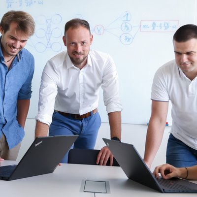 Fünf Menschen befinden sich an einem weißen Tisch. Ganz links sitzt eine Frau an einem Laptop, ganz rechts sitzt ein Mann an einem Laptop. Dazwischen stehen drei Männer. Der Mann in der Mitte blickt in die Kamera, die zwei Personen links blicken auf den linken Laptop, die zwei Personen rechts blicken auf den rechten Laptop.