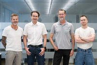 Vier Personen stehen nebeneinander in einem modernen Büroflur mit Glaswänden; sie tragen legere bis business‑casual Kleidung und blicken zur Kamera.