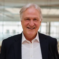 Porträtfoto von Rektor Peter Moser, der lächelnd in die Kamera blickt, gekleidet in einem dunklen Anzug mit weißem Hemd, vor dem Hintergrund des Seminarraums im Studienzentrum.