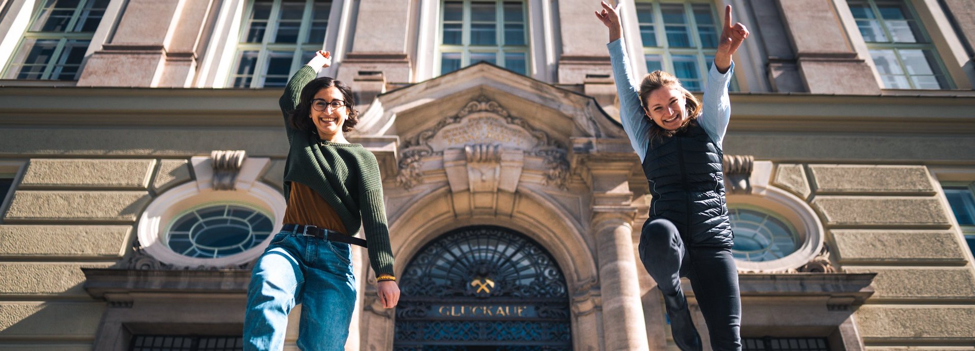 Zwei Studentinnen springen glücklich vor dem Haupteingang der Montanuniversität Leoben in die Luft.