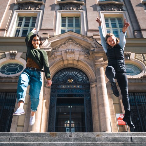 Foto: © MUL Zwei Studentinnen springen lachend vor dem Hauptgebäude der Montanuniversität in die Luft.