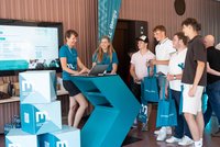 Besucher stehen an einem Info-Stand: Zwei Mitarbeitende in türkisfarbenen Shirts beraten am modernen Thekenpult mit Laptops; Monitor zeigt Programm zum Info‑Tag, daneben gebrandete Würfel und Goodie‑Bags.