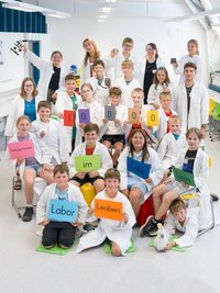Schulklasse und Betreuer:innen in weißen Laborkitteln posieren im Labor und halten bunte Schilder: „10.000 Schüler*innen im Lehr‑Lern‑Labor Leoben".