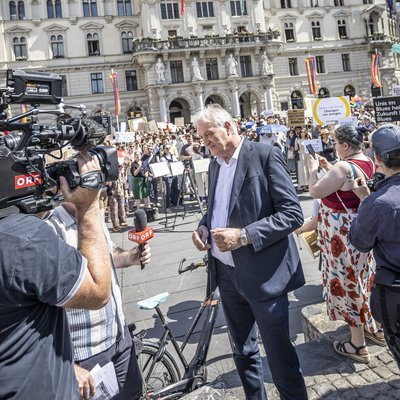 Presse‑ und Publikumsandrang auf einem Platz vor einem prächtigen historischen Rathaus. Kamerateams und Fotografen filmen, Menschen mit Transparenten und Fahnen stehen im Sonnenschein; vorne eine Person im Anzug, im Hintergrund die reich verzierte Fassade mit Turm.