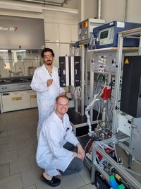 In einem Chemielabor stehen zwei Forschende in Laborkitteln und Schutzbrillen neben einer komplexen Reaktor‑/Messanlage mit Schläuchen, Sensoren und Steuergeräten.