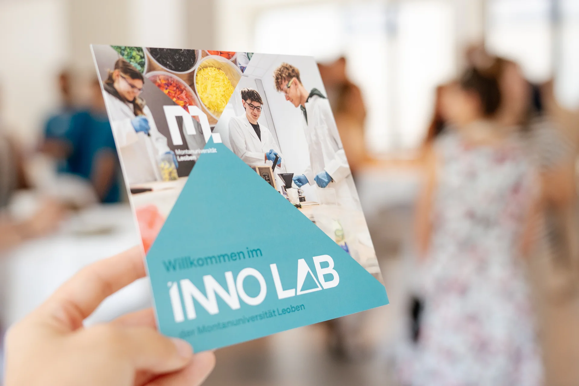 Hand hält einen Flyer „Willkommen im INNO LAB der Montanuniversität Leoben“; im Hintergrund unscharf eine Gruppe bei einer Veranstaltung.