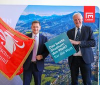 Zwei Männer im Anzug posieren vor einem Panorama‑Hintergrund mit Berg‑/Stadtansicht und Leoben‑Logo. Links wird eine rote Fahne „Österreichischer Städtebund“ gehalten. Rechts halten sie ein Schild mit der Aufschrift „Das beste Studium der Welt? für die Welt“. Gesichter sind unkenntlich gemacht.