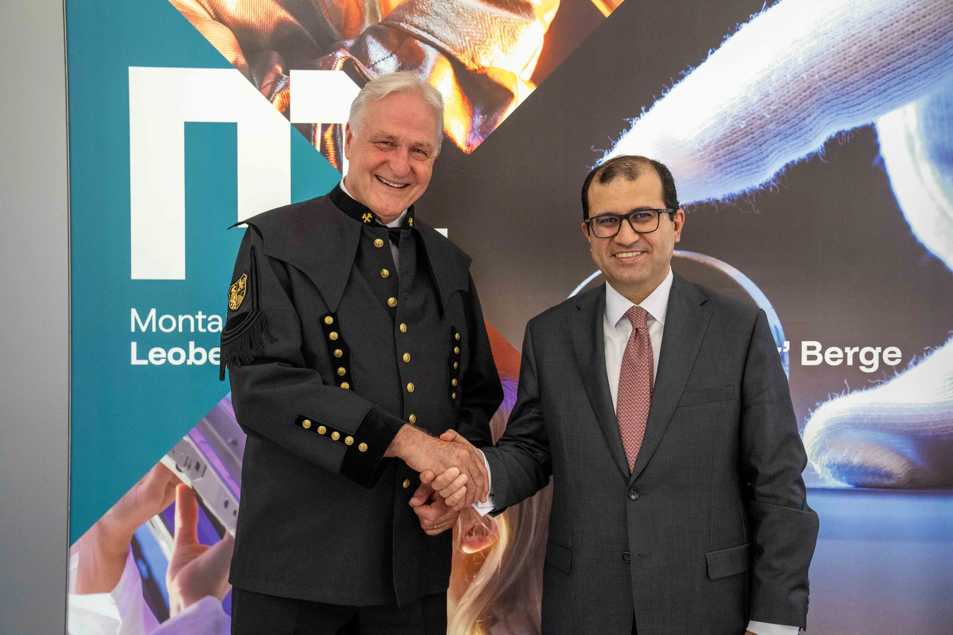 Handshake between two men in front of a university backdrop with logos and imagery. The man on the left wears a dark, historical-style uniform with gold buttons; the man on the right wears a grey suit with a tie. Both are smiling.