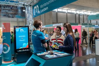 Foto: © MUL In einer Messehalle sprechen drei Besucherinnen an einem blau-türkisen Stand der Montanuniversität Leoben; ein Display zeigt Infos und Broschüren liegen aus.