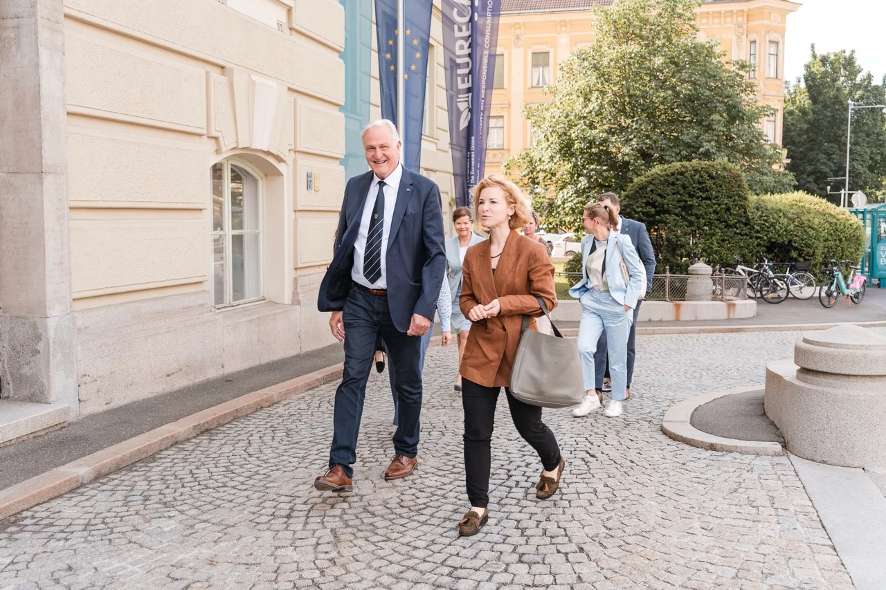 Gruppe in Businesskleidung geht über einen gepflasterten Platz vor einem historischen Gebäude; zwei Personen vorne, weitere folgen. Im Hintergrund Fahnen, Bäume und abgestellte Fahrräder.