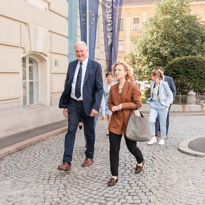 Gruppe in Businesskleidung geht über einen gepflasterten Platz vor einem historischen Gebäude; zwei Personen vorne, weitere folgen. Im Hintergrund Fahnen, Bäume und abgestellte Fahrräder.