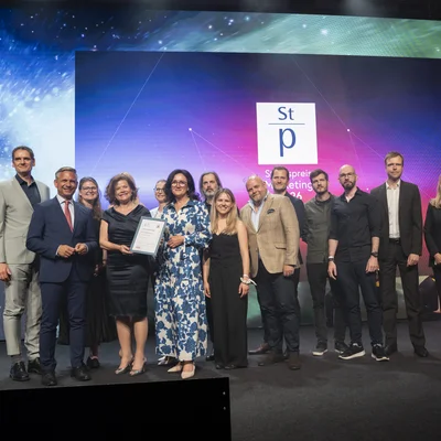 Eine Gruppe von etwa fünfzehn Personen posiert auf einer Bühne bei einer Preisverleihung; eine Person hält eine Urkunde. Hinter ihnen leuchtet eine große LED‑Wand mit farbigem Verlauf und dem Logo „St p“ sowie teilweise lesbarem Text „…preis … 2024“. Alle tragen formelle Kleidung, Bühnenlicht und einige Zuschauer sind im Hintergrund zu sehen.