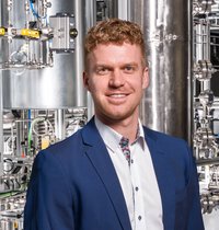Person in blauem Anzug und weißem Hemd vor einer technischen Anlage mit Edelstahlrohren, Ventilen und Messinstrumenten – industrielles/pilotanlagenartiges Umfeld.