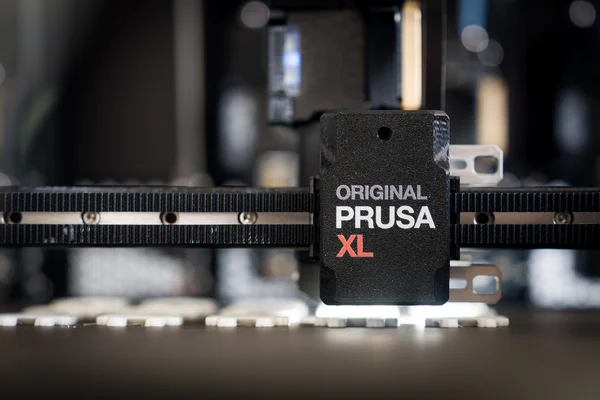 Nahaufnahme eines 3D‑Drucker‑Druckkopfs mit der Aufschrift „Original Prusa XL“ auf einer riemengetriebenen Achse.
