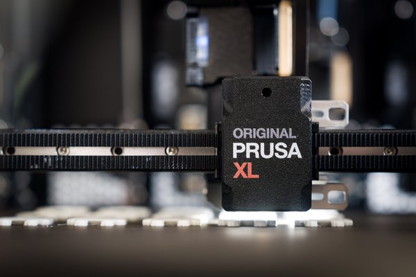 Nahaufnahme eines 3D‑Drucker‑Druckkopfs mit der Aufschrift „Original Prusa XL“ auf einer riemengetriebenen Achse.
