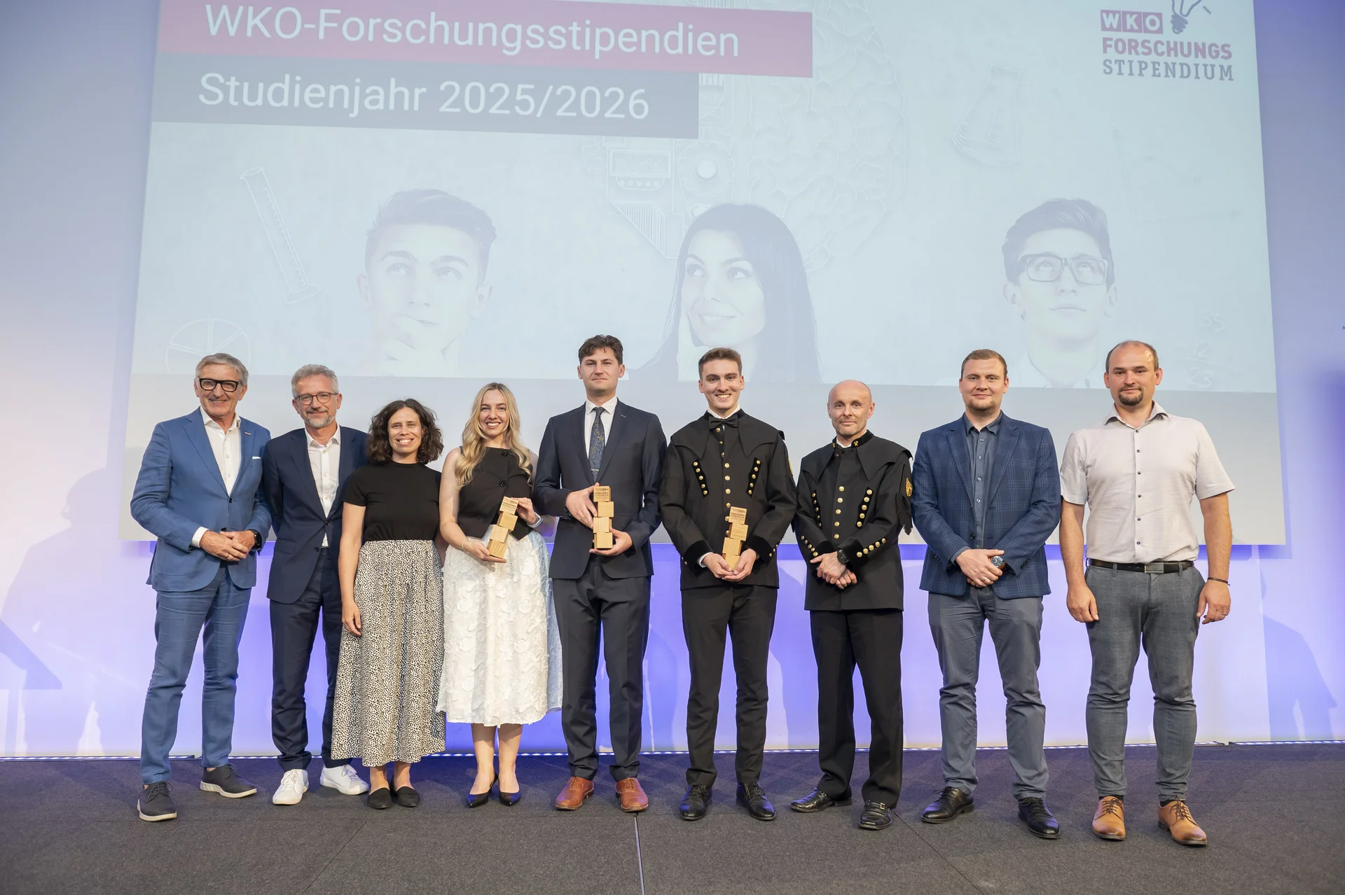 Gruppenfoto auf der Bühne bei der Verleihung der WKO‑Forschungsstipendien 2025/2026: Drei Preisträger*innen mit Trophäen, umgeben von Vertreter*innen.