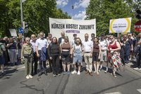 Eine Gruppe von Demonstrierenden steht auf einer Straße vor einem großen Banner mit der Aufschrift „Kürzen kostet Zukunft – #unisretten“. Rechts hält jemand ein Schild „Mehr Bildung, mehr Wissen“. Viele weitere Teilnehmende im Hintergrund, sommerliches Wetter mit Bäumen und blauem Himmel.