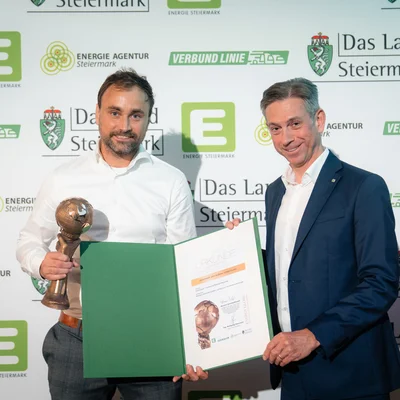 Zwei Personen stehen vor einer Sponsorenwand mit Logos wie „Das Land Steiermark“, „Energie Agentur Steiermark“ und „Verbund Linie“. Die Person links hält eine bronzefarbene Trophäe und ein geöffnetes grün-weißes Zertifikat; die Person rechts trägt einen Anzug – Szene einer Preisverleihung.