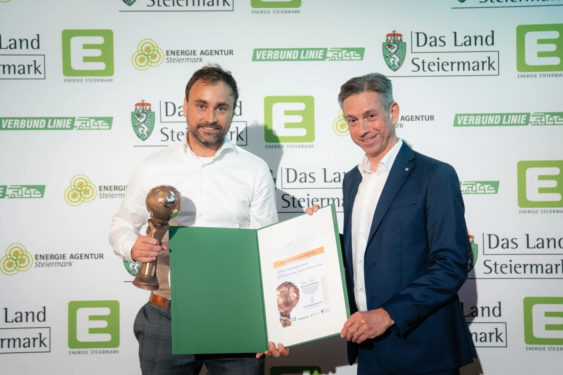 Zwei Personen stehen vor einer Sponsorenwand mit Logos wie „Das Land Steiermark“, „Energie Agentur Steiermark“ und „Verbund Linie“. Die Person links hält eine bronzefarbene Trophäe und ein geöffnetes grün-weißes Zertifikat; die Person rechts trägt einen Anzug – Szene einer Preisverleihung.