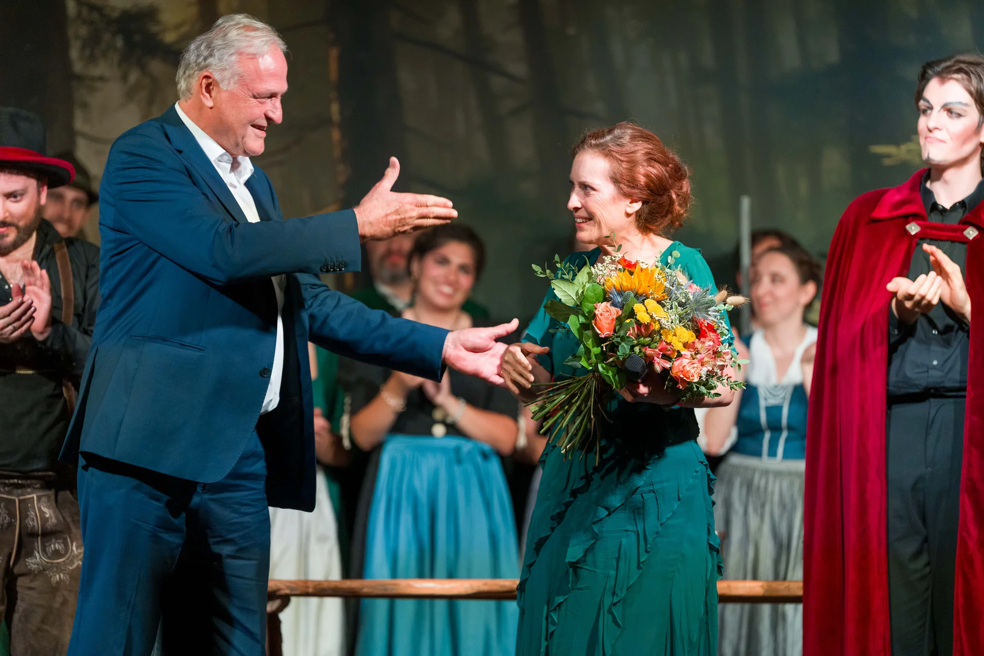 Bühnenmoment nach der Aufführung: Eine Person im Anzug gratuliert einer Frau in grünem Kleid, die einen großen Blumenstrauß hält; Ensemblemitglieder in Kostümen applaudieren im Hintergrund.