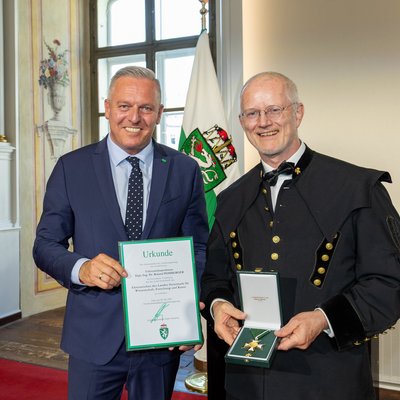 Verleihung des Ehrenzeichens des Landes Steiermark für Wissenschaft, Forschung und Kunst: Person im Anzug hält die Urkunde, daneben Person in Bergmannsuniform präsentiert den Orden; Flagge im historischen Saal.