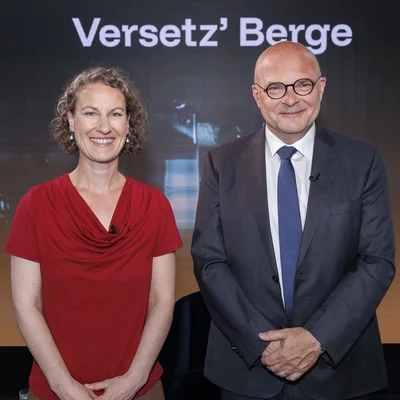      Zwei Personen vor einer Bühnenwand mit „Versetz’ Berge“ und Logo der Montanuniversität Leoben; eine trägt Rot, die andere Anzug und Krawatte.