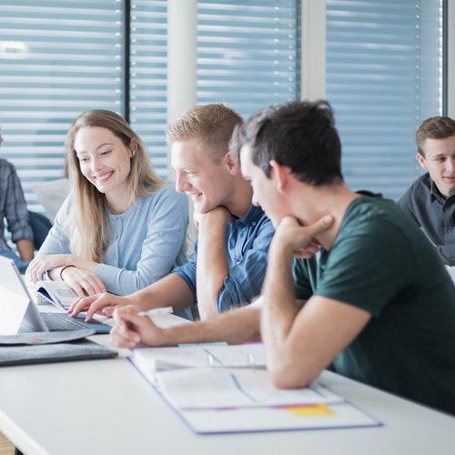 Studenten und Studentinnen sitzen in einem Seminarraum am Tisch und sehen gemeinsam auf einen Laptop.