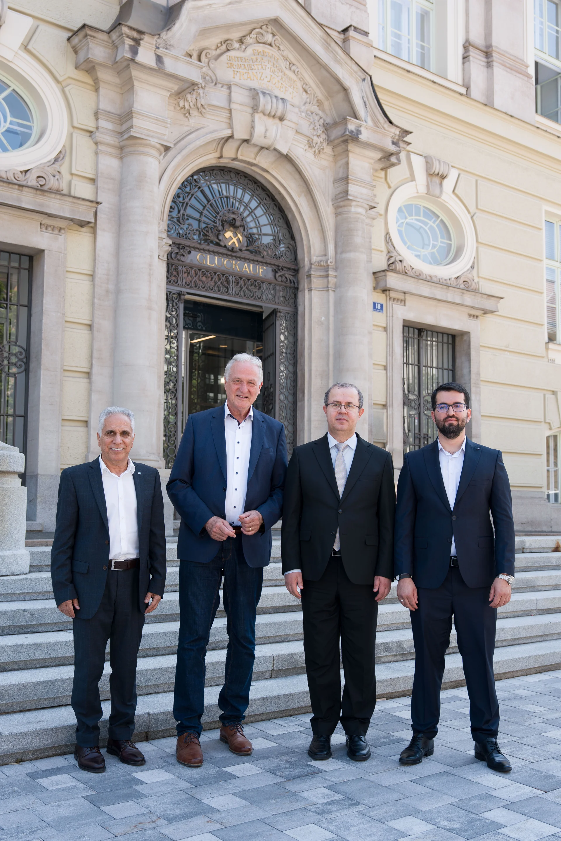 Vier Personen in formeller Kleidung stehen für ein Gruppenfoto vor einem historischen Gebäudeeingang.