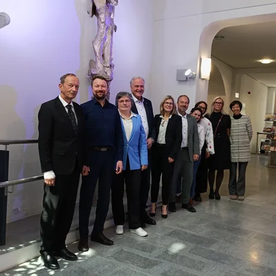 Gruppenfoto in einem hellen Foyer: Mehrere Personen in Business‑Kleidung stehen nebeneinander und posieren. Links an der Wand hängt eine Skulptur; rechts führen Bögen in einen Empfangsbereich mit Theke und Glas­türen. Polierter Steinboden, Tageslicht.