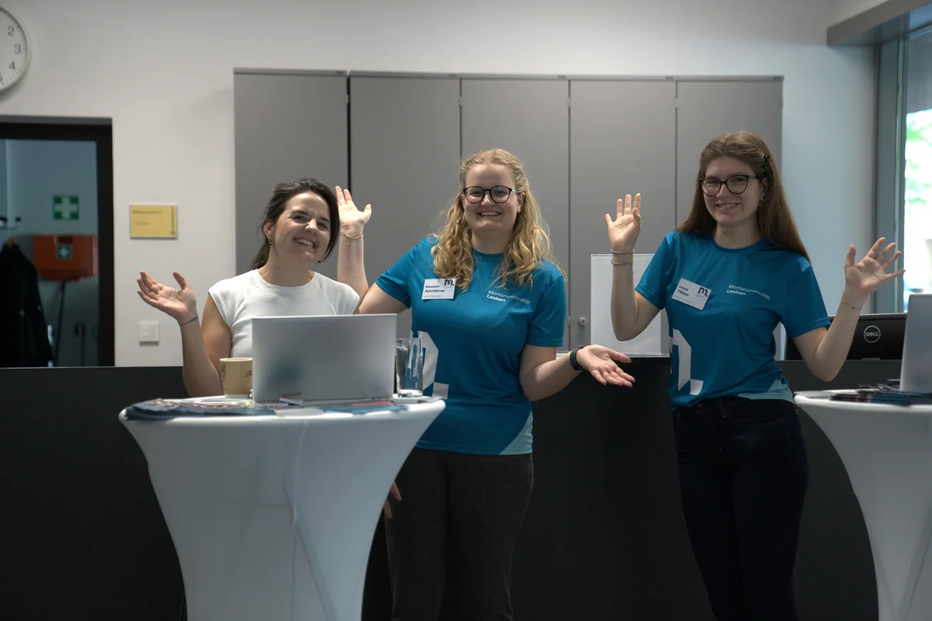 Drei Studienberaterinnen warten fröhlich und mit in die Luft gestreckten Armen auf Studieninteressierte.