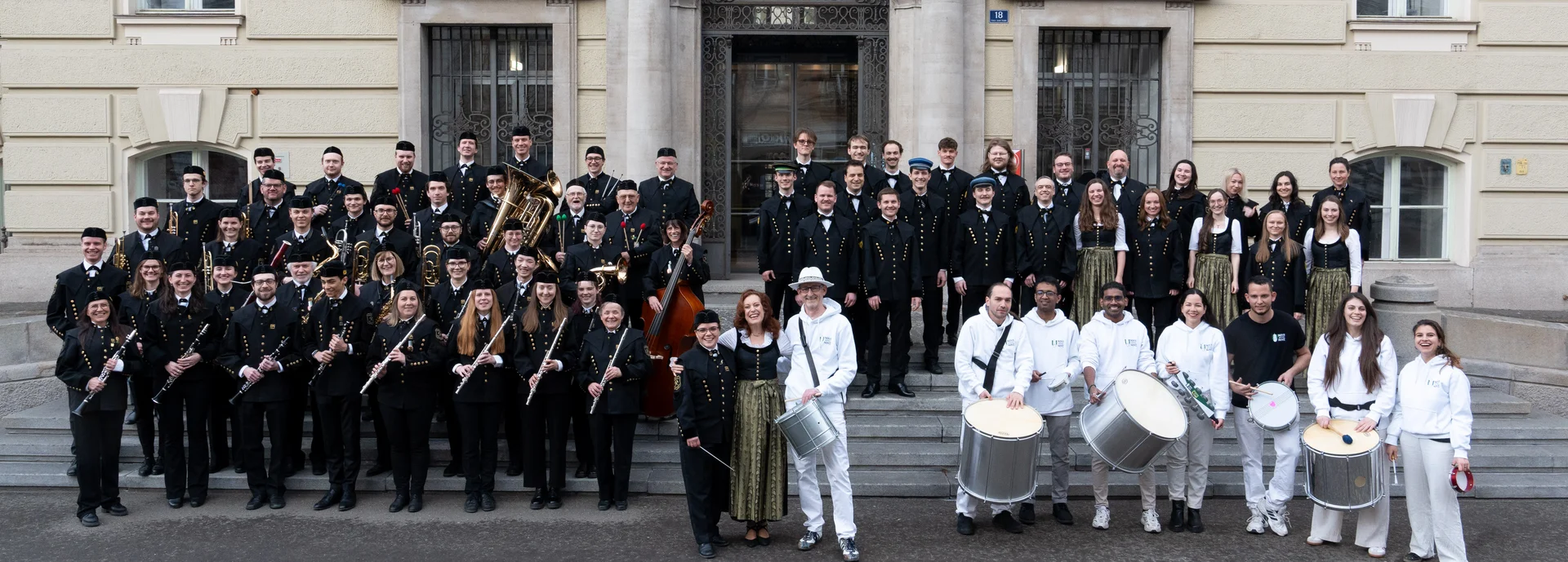 The members of the university wind orchestra, the university choir and the drum formation stand together on the outside steps of the main building.