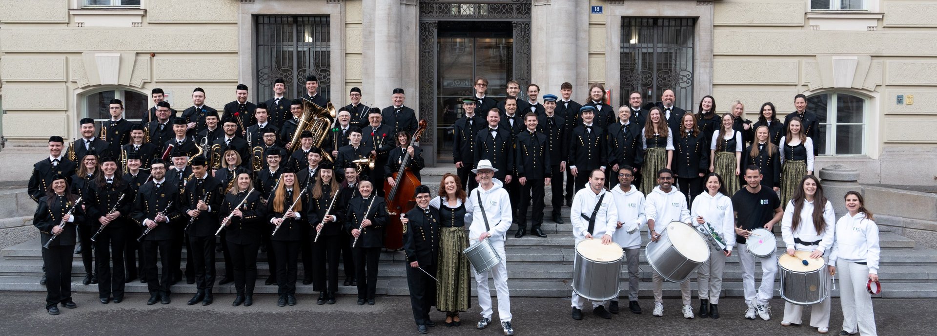 The members of the university wind orchestra, the university choir and the drum formation stand together on the outside steps of the main building.