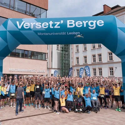 Große Laufgruppe unter einem türkisfarbenen Startbogen mit „Versetz’ Berge – Montanuniversität Leoben“. Viele heben die Arme; gelbe und blaue Event‑Shirts, Campus- und Altstadtgebäude im Hintergrund.