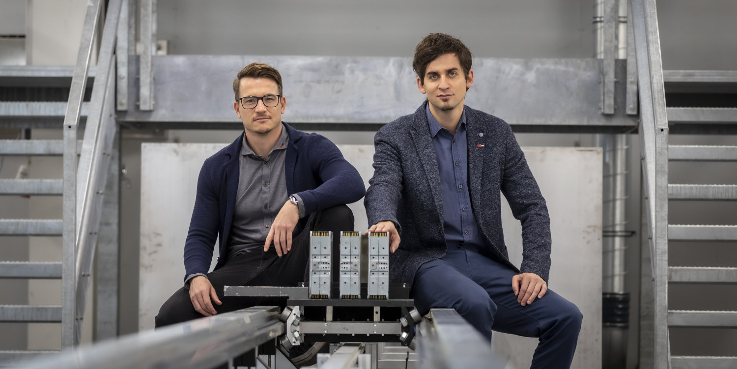 Jörg Moser (links) und Christian Ellersdorfer vom Institut für Fahrzeugsicherheit der TU Graz an einem Prüfstand des Battery Safety Centers Graz. Bildquelle: Lunghammer - TU Graz Zwei Männer sitzen auf den Schienen eines Prüfstandes und schauen in die Kamera