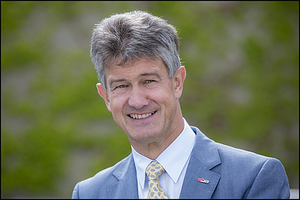 Rektor Harald Kainz mit grauem Sakko und gemusterter Krawatte vor grünem verschwommenem Hintergrund.