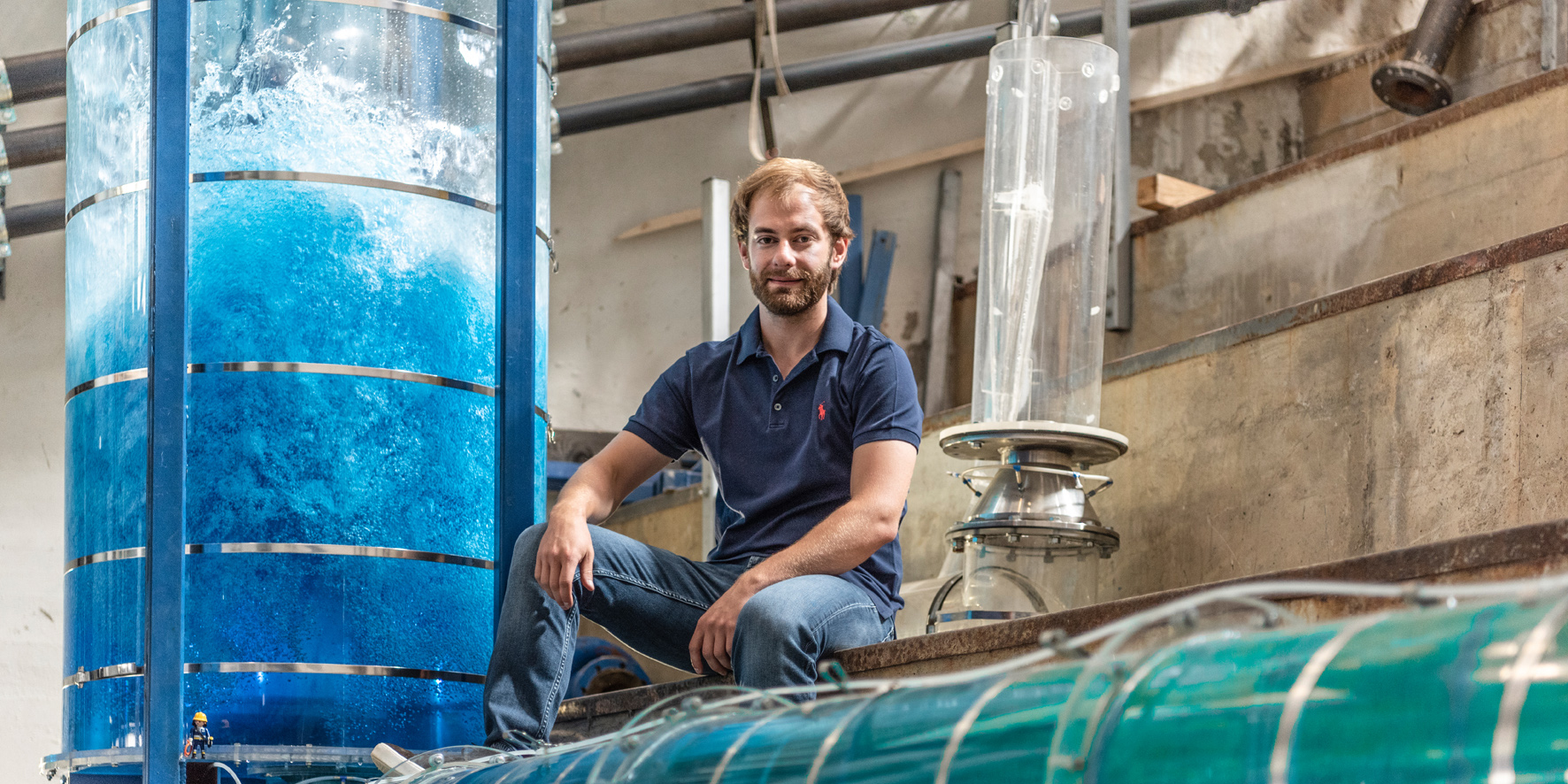 Ein junger Mann mit Bart in Jeans und blauem T-Shirt sitzt im Wasserbaulabor und ist umgeben von Versuchanordnungen in Form von wassergefüllten Plexiglas-Röhren.